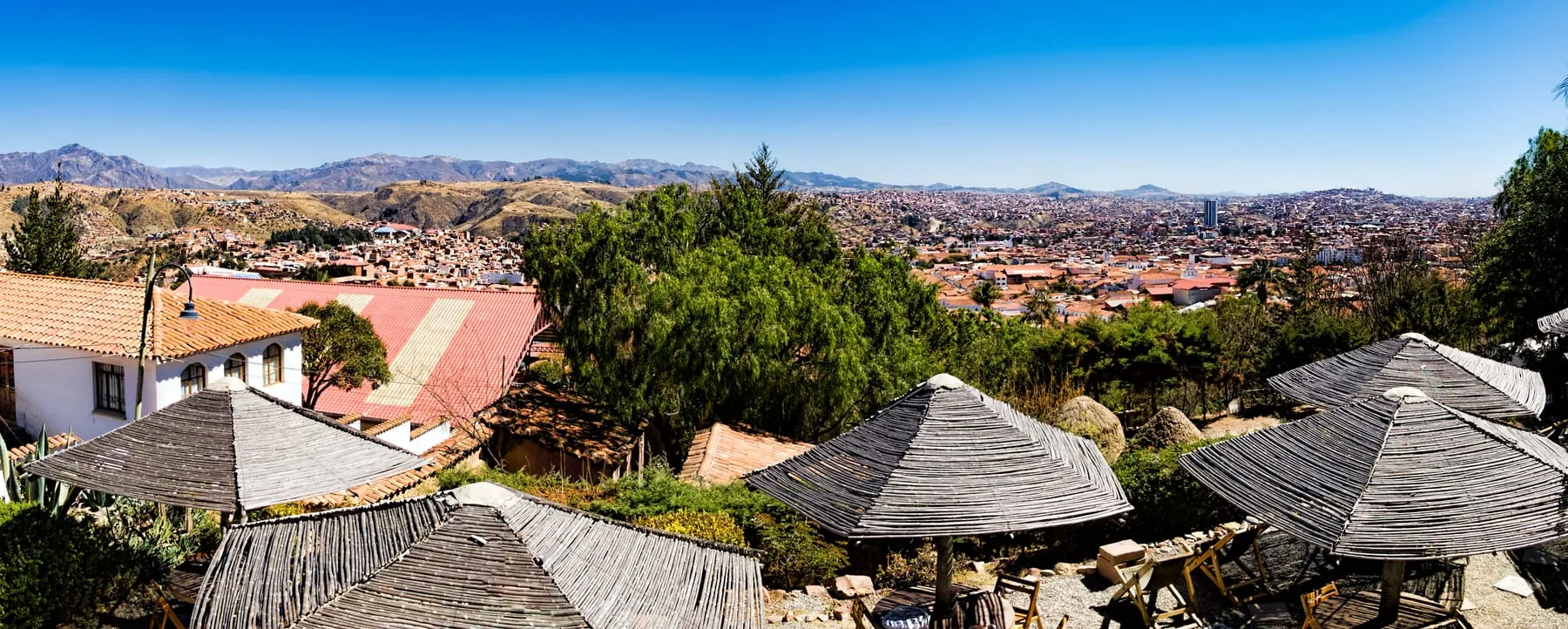 Panorámica amplia de la ciudad de Sucre extendida en un valle rodeado de cerros, con techos de teja roja y casas blancas.
