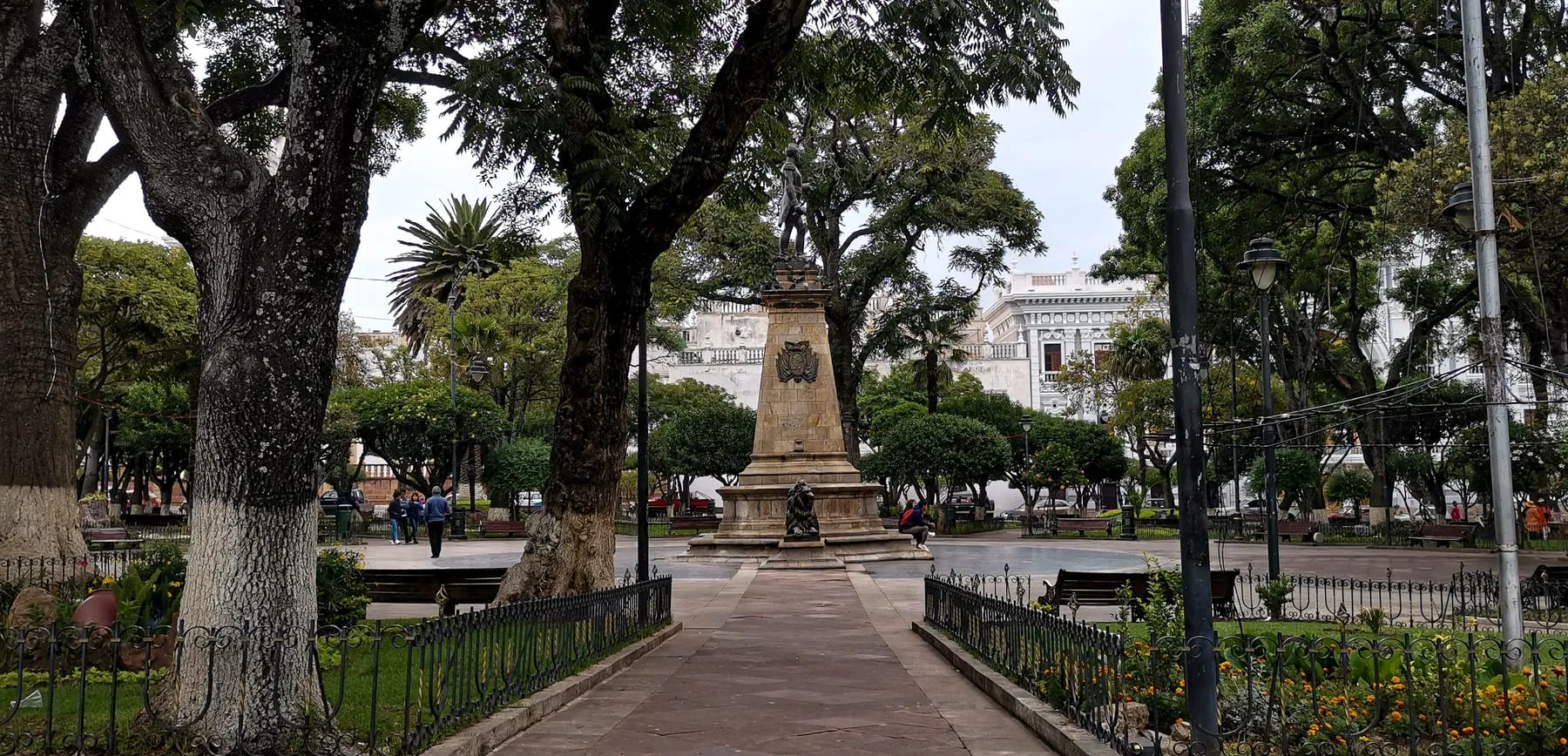 Vista de la Plaza 25 de Mayo de Sucre con árboles frondosos, senderos empedrados, bancos y el monumento central sobre un pedestal de piedra.