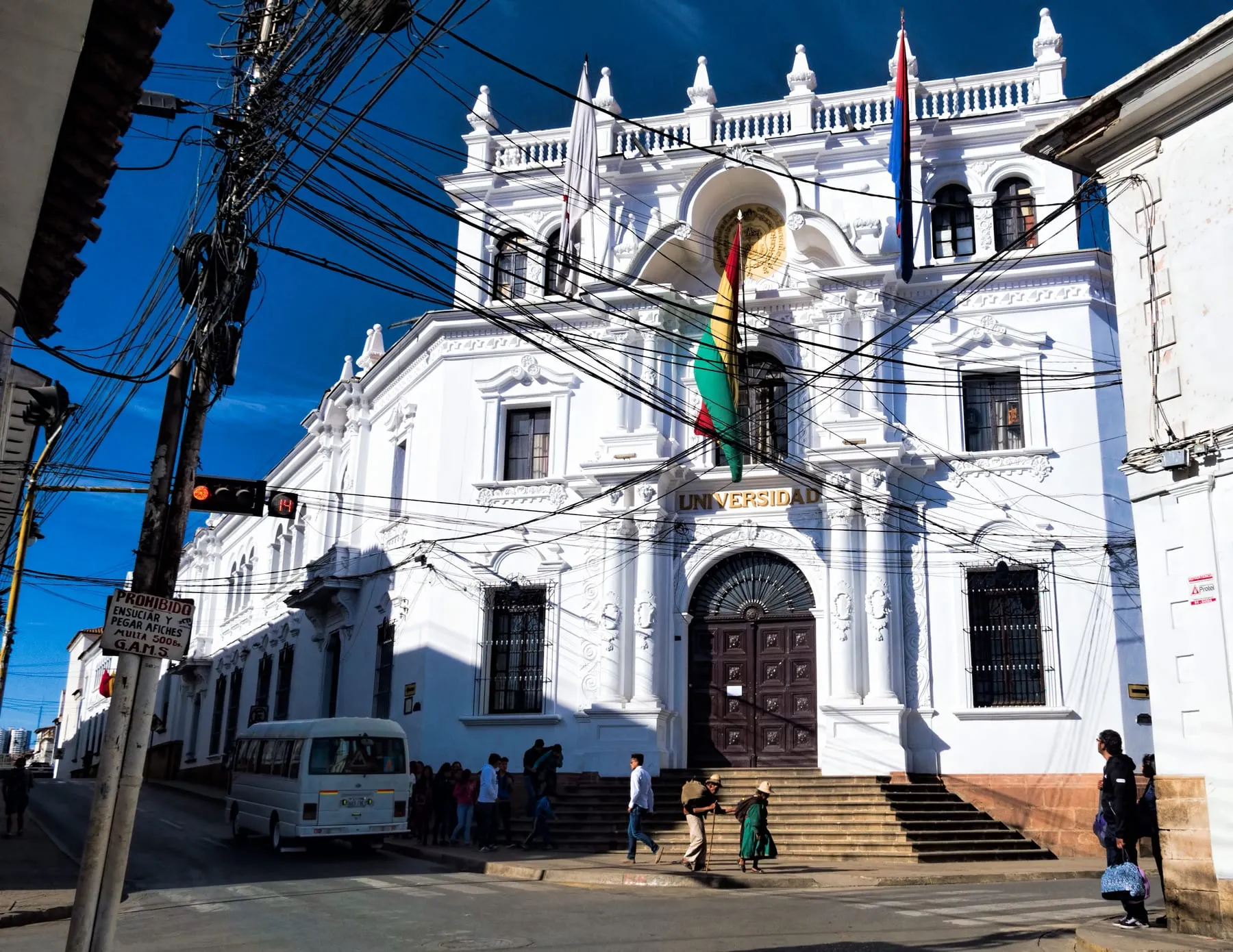Fachada blanca de estilo colonial del edificio central de la Universidad de San Francisco Xavier en Sucre, con banderas de Bolivia y Chuquisaca y cielo azul intenso.