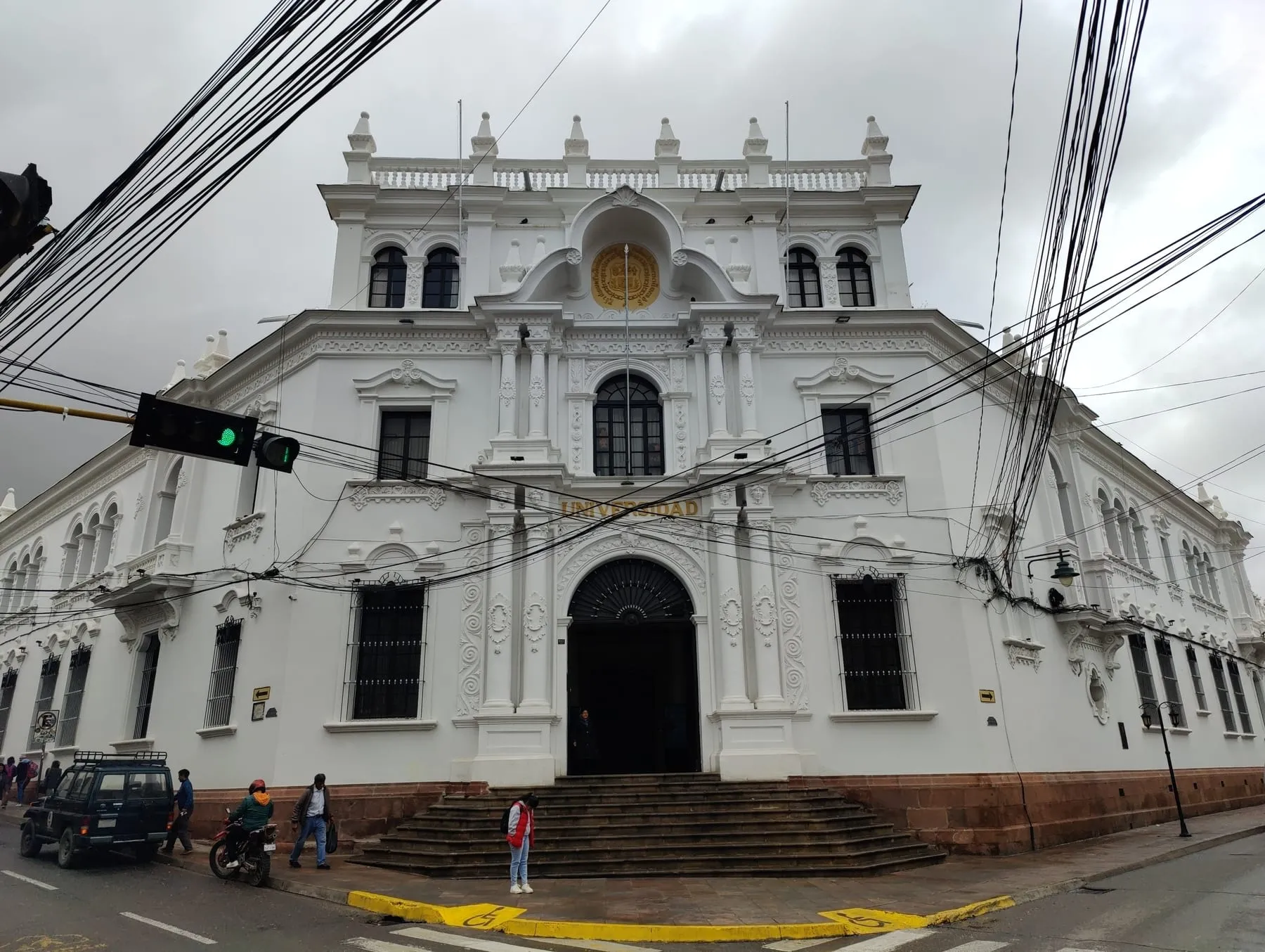 Vista angular del edificio blanco de la Universidad de San Francisco Xavier en una esquina de Sucre, con escalinata de piedra, arco de entrada y escudo dorado en el frontón.
