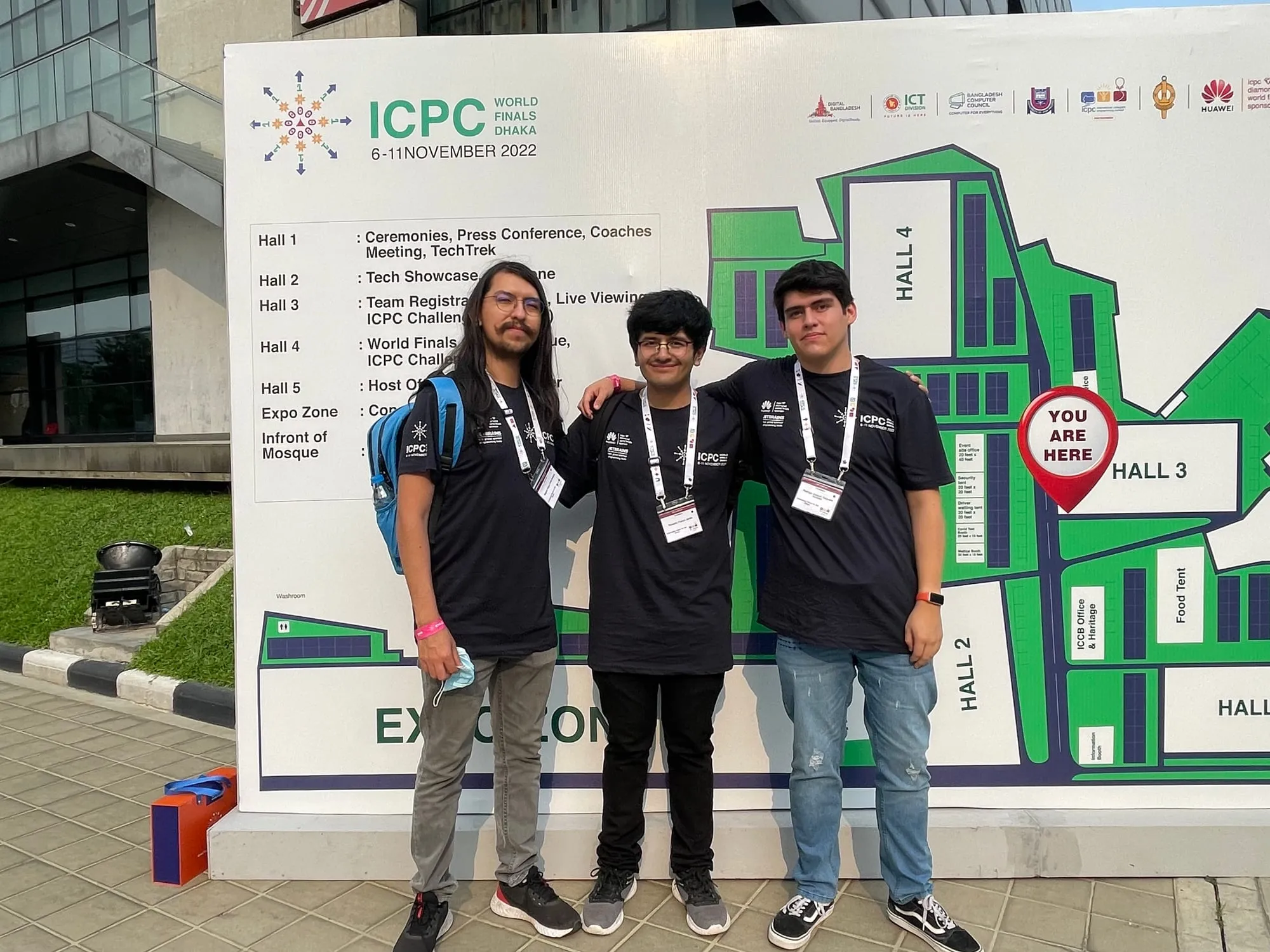 Tres integrantes del equipo boliviano posando frente al cartel oficial de las Finales Mundiales del ICPC en Dhaka.